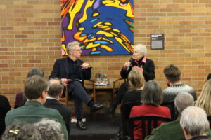 Author Margaret Atwood speaks into a microphone while seated onstage beside moderator James Orbinski during a public discussion at Massey College. They sit in front of a colourful painting and a brick wall, facing an audience filling the room.