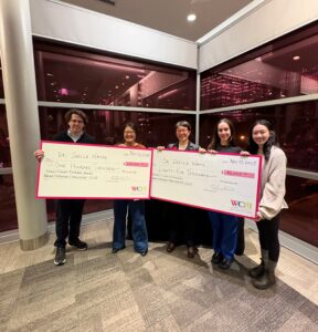 Six people hold two oversized cheques for $100,000 and $85,000, smiling indoors at night with city lights behind them.